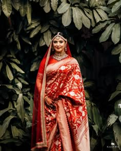 a woman in a red and white sari standing next to green leaves with her hands on her hips