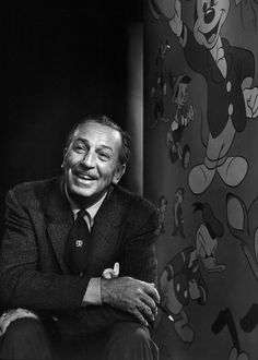 a man in a suit and tie sitting next to a mickey mouse wall with his hands on his knees