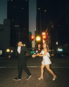 a man and woman dancing in the middle of a city street at night with buildings lit up behind them