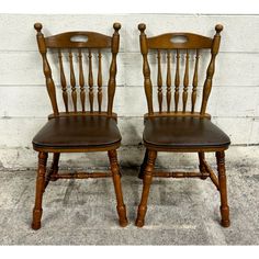 two wooden chairs sitting side by side against a white brick wall, one with brown leather seat covers