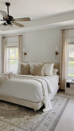 a large white bed sitting inside of a bedroom next to two windows and a ceiling fan