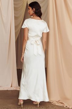 a woman in a white dress standing next to a curtain and looking off into the distance