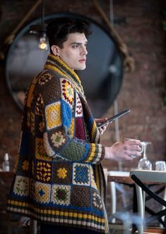 a man standing in front of a table holding a cell phone and wearing a crocheted jacket