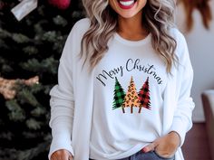 a woman standing in front of a christmas tree wearing a white sweatshirt with merry christmas trees on it