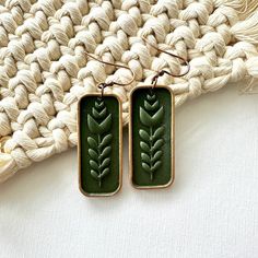 pair of earrings with green leaves on them sitting on top of a woven piece of cloth