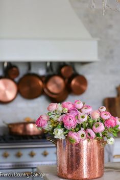 pink and white flowers in a copper vase on a kitchen counter top with copper pots hanging above