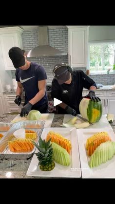 two men in kitchen preparing food on trays and cutting up melons with knifes