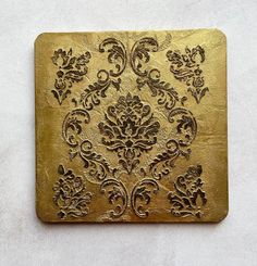 a gold square with an ornate design on the front and back side, sitting on a white surface