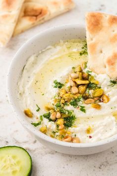 a white bowl filled with hummus and pita bread next to cucumber