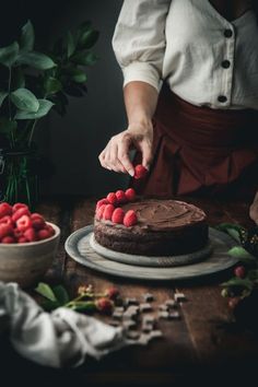 a woman is decorating a chocolate cake with raspberries on the table in front of her