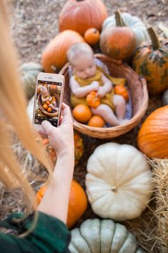 a woman taking a photo of a baby in a basket surrounded by pumpkins and gourds