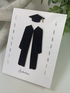 a graduation card with a graduate's cap and gown cut out on it, sitting next to a plant