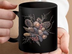 a person holding a black coffee mug with pine cones and berries