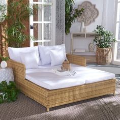 a wicker daybed with white pillows in front of a window and potted plants