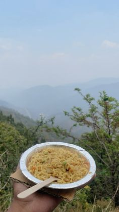 a hand holding a paper plate with noodles in it and mountains in the back ground