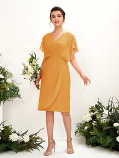 a woman standing in front of flowers and greenery wearing a yellow dress with short sleeves