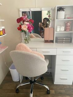 a white desk with a mirror and flowers on it in front of a shelf filled with other items