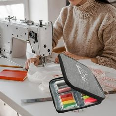 a woman sitting at a sewing machine with markers in front of her and an open case on the table