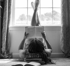 a woman laying on the floor reading a book with her legs up in front of an open window