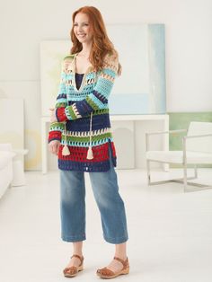 a woman standing in front of a white wall wearing jeans and a colorful cardigan