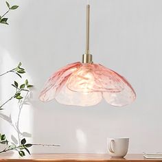 a pink flower shaped light hanging over a wooden table next to a white cup and plant