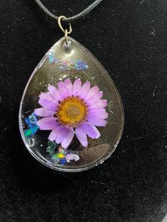 a purple flower in a tear shaped glass pendant on a black surface with a silver chain