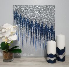 two candles are next to a painting with blue and silver glitters on it, along with a potted plant