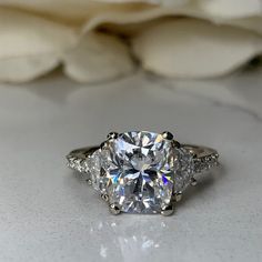 a close up of a diamond ring on a white surface with flowers in the background