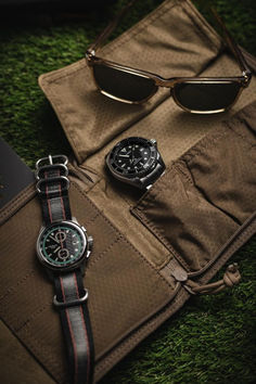 two watches sitting on top of a brown bag next to some sunglasses and a book