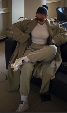 a woman sitting on top of a black couch wearing white sneakers and a trench coat