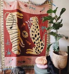 two cats sitting on the floor next to a potted plant and a wall hanging