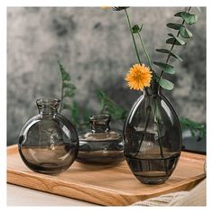 two clear vases with yellow flowers in them on a wooden tray next to a gray wall