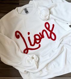 Pullover embroidered with cursive chenille Lions Team. Will be made as pictured. Please message us for custom colors, however we do have most basic colors of chenille available. *We use only the highest quality Gildan Heavy Blend Unisex Sweatshirts White T-shirt With Embroidered Text For Fall, White Embroidered Logo Top For College, White Cotton Varsity Sweater, Oversized Embroidered White Sweater, Oversized Embroidered White Top, Oversized White Embroidered Top, College Red Tops With Embroidered Text, White Sweatshirt With Custom Embroidery, Relaxed Fit, White Sweatshirt With Custom Embroidery In Relaxed Fit