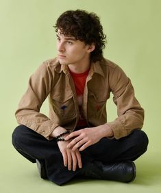 a young man sitting on the ground with his hands in his pockets, wearing a brown shirt and black pants