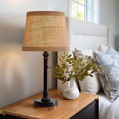 a lamp on a night stand next to a bed with white pillows and throw pillows