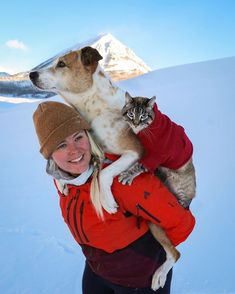 a woman holding a cat and dog on her back in the snow with mountains in the background