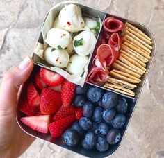 a hand holding a bento box filled with fruit and crackers, cheeses, strawberries