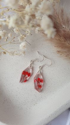 two red earrings sitting on top of a white table next to dried flowers and feathers