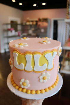 a hand holding a pink and yellow cake with white flowers on the top, in front of a store display