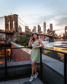 a woman in a dress standing on a balcony next to the brooklyn bridge at sunset