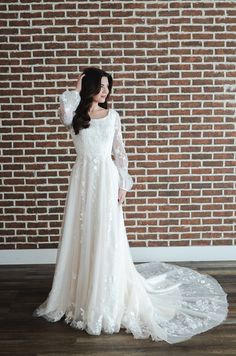 a woman standing in front of a brick wall wearing a wedding dress with long sleeves