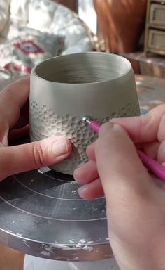 two hands are working on a vase with a pink pen in front of the cup