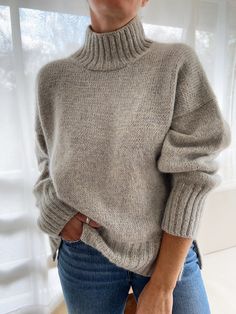 a woman standing in front of a window wearing a gray sweater and jeans with her hands on her hips