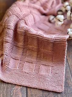 a pink knitted blanket sitting on top of a wooden floor next to cotton balls