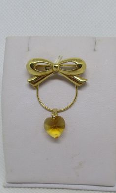 Never Worn Vintage Trifari gold-tone metal bow wire hanger yellow faceted crystal heart pendant charm holder badge hanger eyeglass holder ID tag holder with a standard C clasp. The wire hanger allows you to slide additional charms onto it. The bow is hallmarked Trifari. Length including bow hanger section & crystal heart 1 & 1/2 inches. Width of bow 1 & 1/8 inches. Width across of wire hanger 3/4 inch. Heart length 7/16 inch. Heart width 7/16 inch. I offer multiple-item-order shipping discounts. Elegant Yellow Brooches For Gifts, Yellow Brooch Jewelry For Wedding, Yellow Wedding Jewelry Brooch, Yellow Gold Pendant Brooches As Gift, Gold Round Brooches For Gifts, Heart-shaped Brooch Jewelry Gift, Elegant Gold Brooch For Valentine's Day, Elegant Gold Brooches For Valentine's Day, Gold Heart-shaped Brooches For Gifts