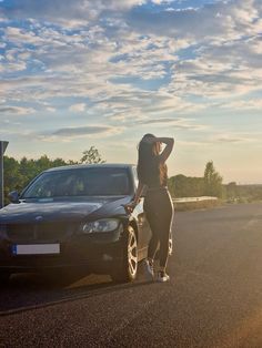 a woman standing next to a car on the side of the road with her hands behind her head