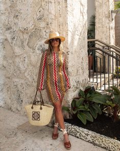 a woman standing next to a wall holding a purse and wearing a straw hat with her hands in her pockets