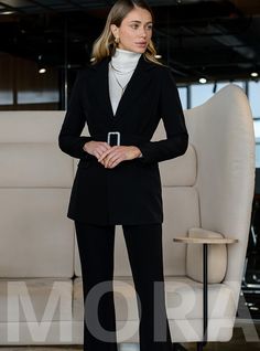 a woman standing in front of a couch wearing a black suit and white turtle neck sweater