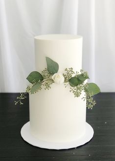 a white wedding cake with greenery and flowers on the top is sitting on a black table