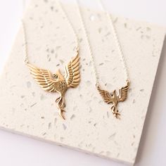 two gold necklaces with birds on them sitting on a white card holder next to each other